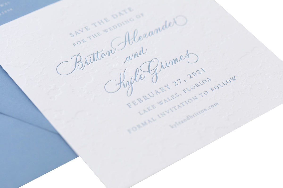 A white letterpress save the date card with elegant blue script announces the wedding of Britton Alexander and Kyle Grimes on February 27, 2021, in Lake Wales, Florida. Personalized paper cards and a blue envelope add to its timeless charm.