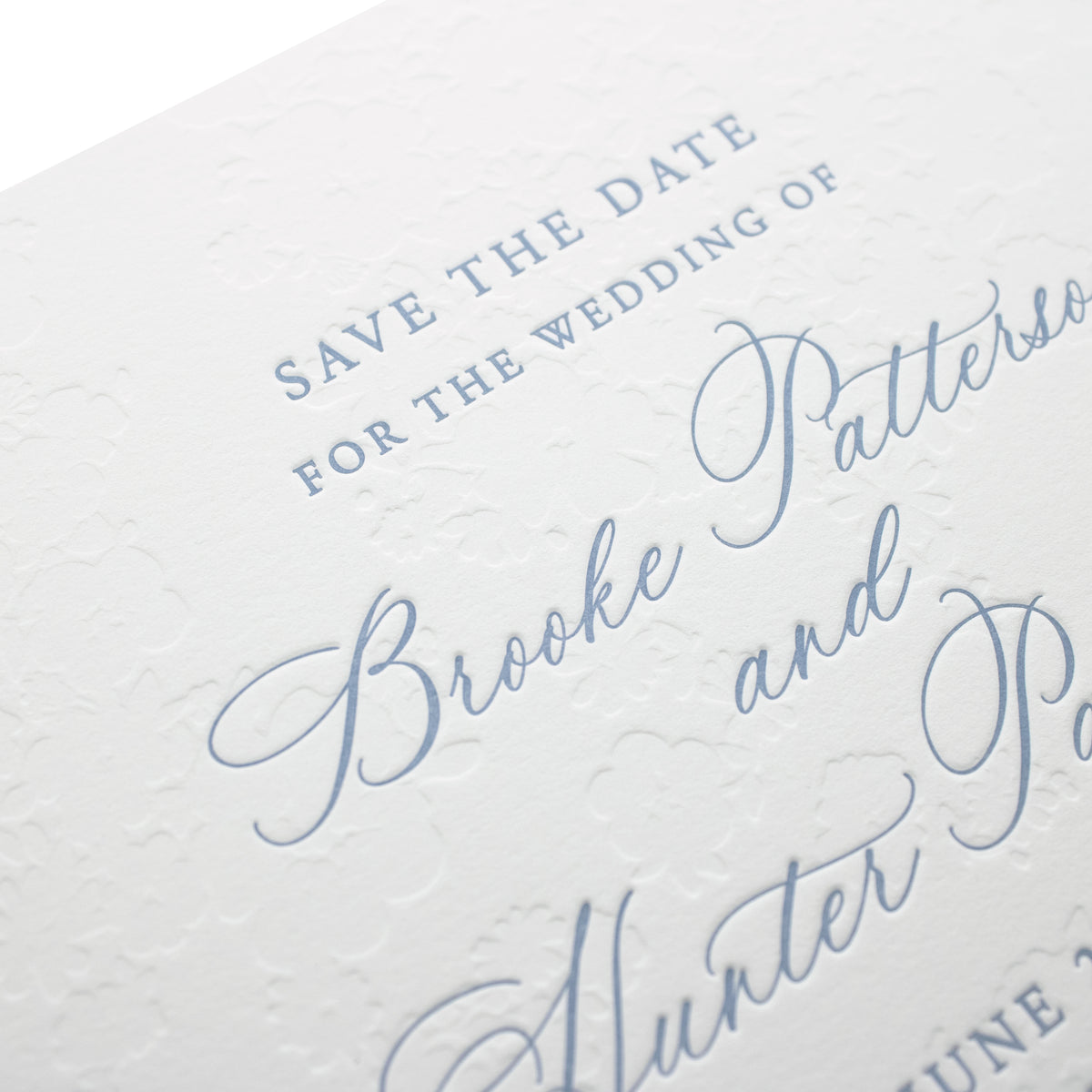Close-up of a white wedding invitation card with elegant blind debossed floral patterns and light blue cursive text, showcasing a personalized paper card announcing the wedding of Brooke Patterson and Hunter Patterson.