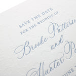 Close-up of a white wedding invitation card with elegant blind debossed floral patterns and light blue cursive text, showcasing a personalized paper card announcing the wedding of Brooke Patterson and Hunter Patterson.