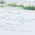 Close-up of a personalized stationery wedding invitation with elegant blue script, event dates (October 2-4, 2020), and Sea Island, Georgia location, featuring a watercolor save the date beach scene at the top.