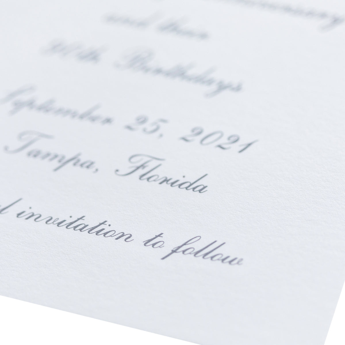 Close-up of an elegant, white textured engraved card with cursive gray text mentioning September 25, 2021, Tampa, Florida, and the phrase invitation to follow. The card is partially shown and softly focused.