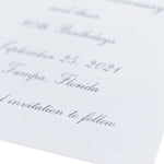 Close-up of an elegant, white textured engraved card with cursive gray text mentioning September 25, 2021, Tampa, Florida, and the phrase invitation to follow. The card is partially shown and softly focused.