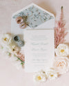 Wedding invitation card with floral design, rings, and flowers on a light pink background