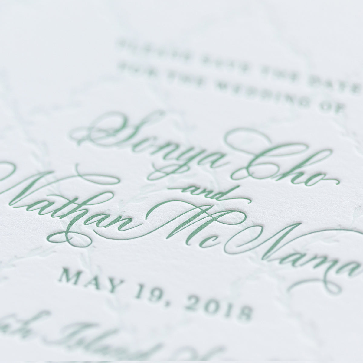 A close-up photo of an elegant wedding invitation on personalized paper with the names Senya Cho and Nathan McNamara in green cursive, and May 19, 2018, below. The subtle textured design highlights exquisite letterpress details.