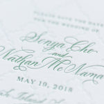 A close-up photo of an elegant wedding invitation on personalized paper with the names Senya Cho and Nathan McNamara in green cursive, and May 19, 2018, below. The subtle textured design highlights exquisite letterpress details.